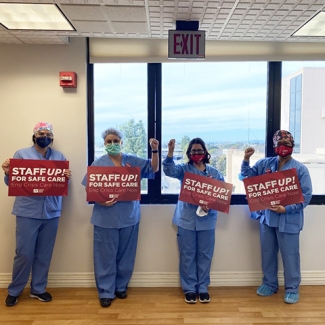 Four nurses inside hospital holding signs "Staff Up for Safe Patient Care"