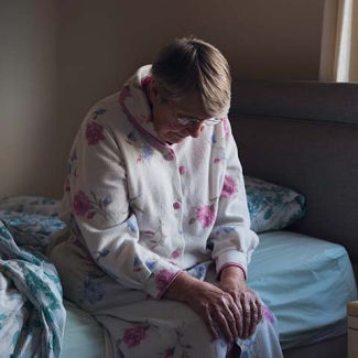 Older woman sits on side of bed with head down
