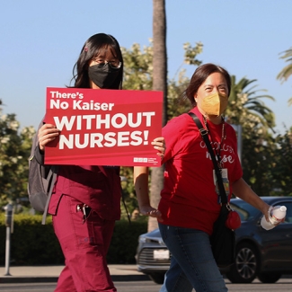 Two nurses outside, one holds sign "There's No Kaiser Without Nurses"