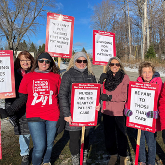 Group of nurses outside dressed in winter clothes, holding signs "Standing for our patients", "Nurses are the heart of patient care", more
