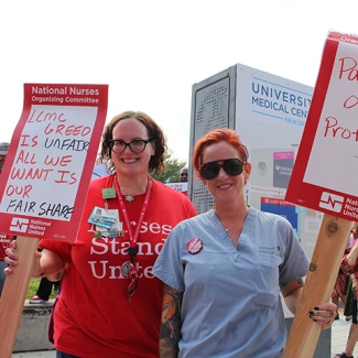 Two nurses smiling, holding signs "LCMC Greed Is Unfair, All We Want Is Our Fair Share" and "Patients Over Profit"