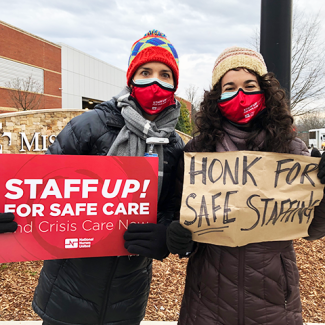 Two nurses outside MIssion Hospital holding signs: "Staff up! for safe care. End crisis care now." and "Honk for safe staffing."