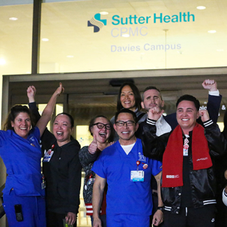 Large group of nurses outside hospital, smiling, with raised fists