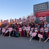 Large group of nurses outside San Joquin General Hospital, smiling and holding signs "RNs On Strike" and "Respect Nurses"