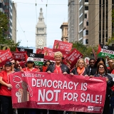 Large group of nurses marching, holding banner "Democracy is NOT for Sale"