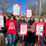 Group of nurses outside dressed in winter clothes, holding signs "Standing for our patients", "Nurses are the heart of patient care", more