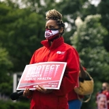 Masked nurse outside holds signs "Protect Nurses, Patients, Public Health"